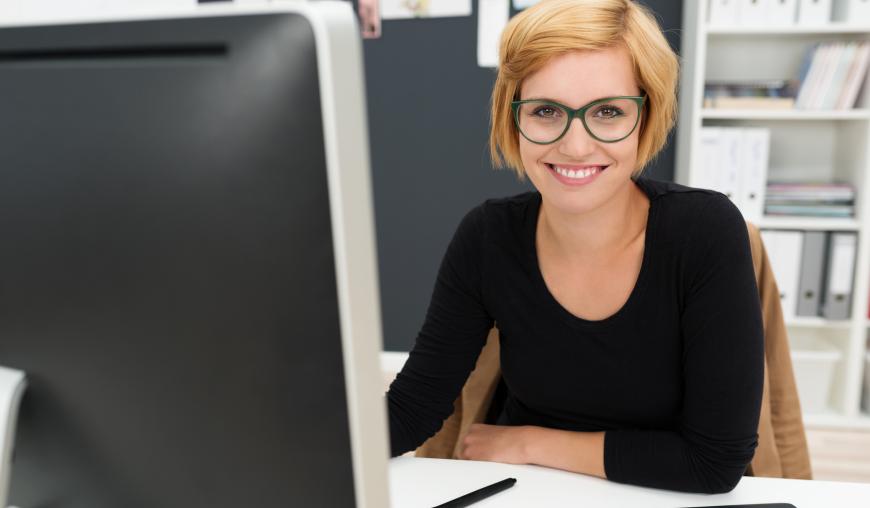 Eine Frau sitzt an einem Schreibtisch vor einem Computer in einem Büro. Sie lächelt freundlich in die Kamera.