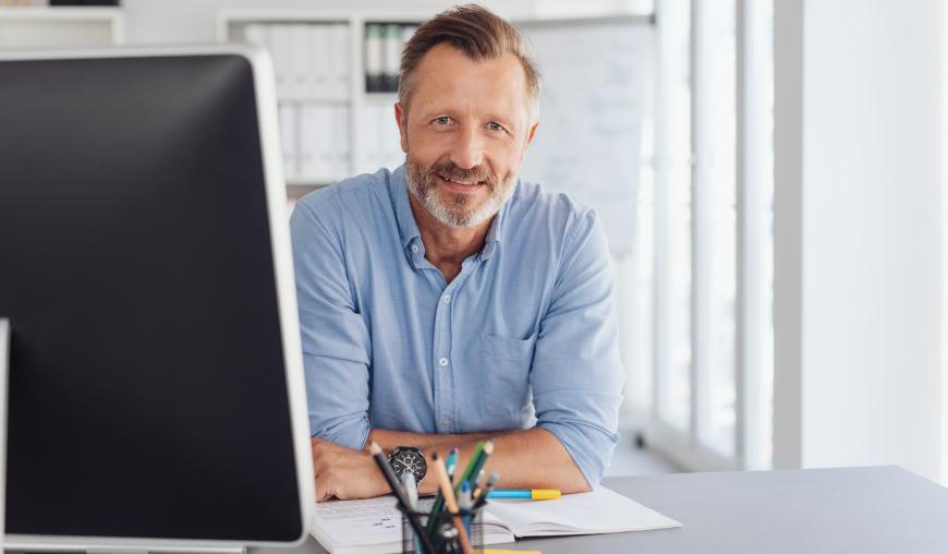 Ein freundlich lächelnder Mann in fortgeschrittenem Alter sitzt an einem Schreibtisch vor einem Computer.