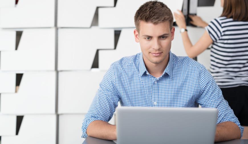 Ein junger Mann sitzt in einem Büro und konzentriert sich auf seinen Laptop. Im Hintergrund holt eine Frau einen Aktenordner aus dem Schrank.