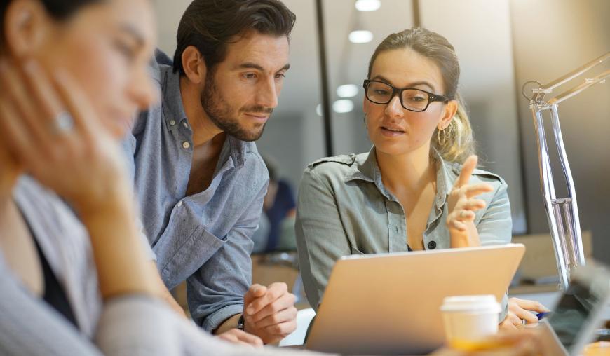 Eine Frau und ein Mann beugen sich gemeinsam über einen Laptop. Sie erklärt ihm etwas. Im Vordergrund sitzt eine weitere Frau mit einem Handy in der Hand.